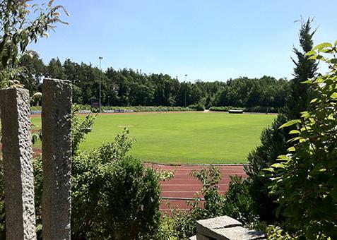 Sportanalage am Kosbacher Weg Sportanlage mit Laufbahn und Rasenfeld, teilweise von Büschen und Steinen im Vordergrund eingerahmt.
