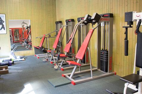 Fitnessgeräte im Kraftraum der Jahnhalle Blick auf Fitnessgeräte im Kraftraum der Jahnhalle, die an der Wand entlang aufgestellt sind, mit einem Spiegel im Hintergrund.