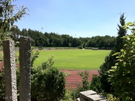 Sportplatz am Kosbacher Weg Sportanlage mit Laufbahn und Rasenfeld, teilweise von Büschen und Steinen im Vordergrund eingerahmt.