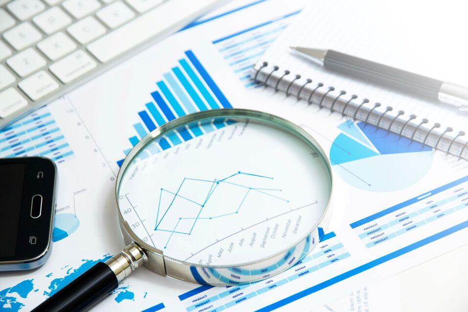 Analyse von Geschäftsdaten Eine Lupe liegt auf Dokumenten mit Diagrammen und Statistiken, daneben ein Smartphone, eine Tastatur und ein Notizbuch.